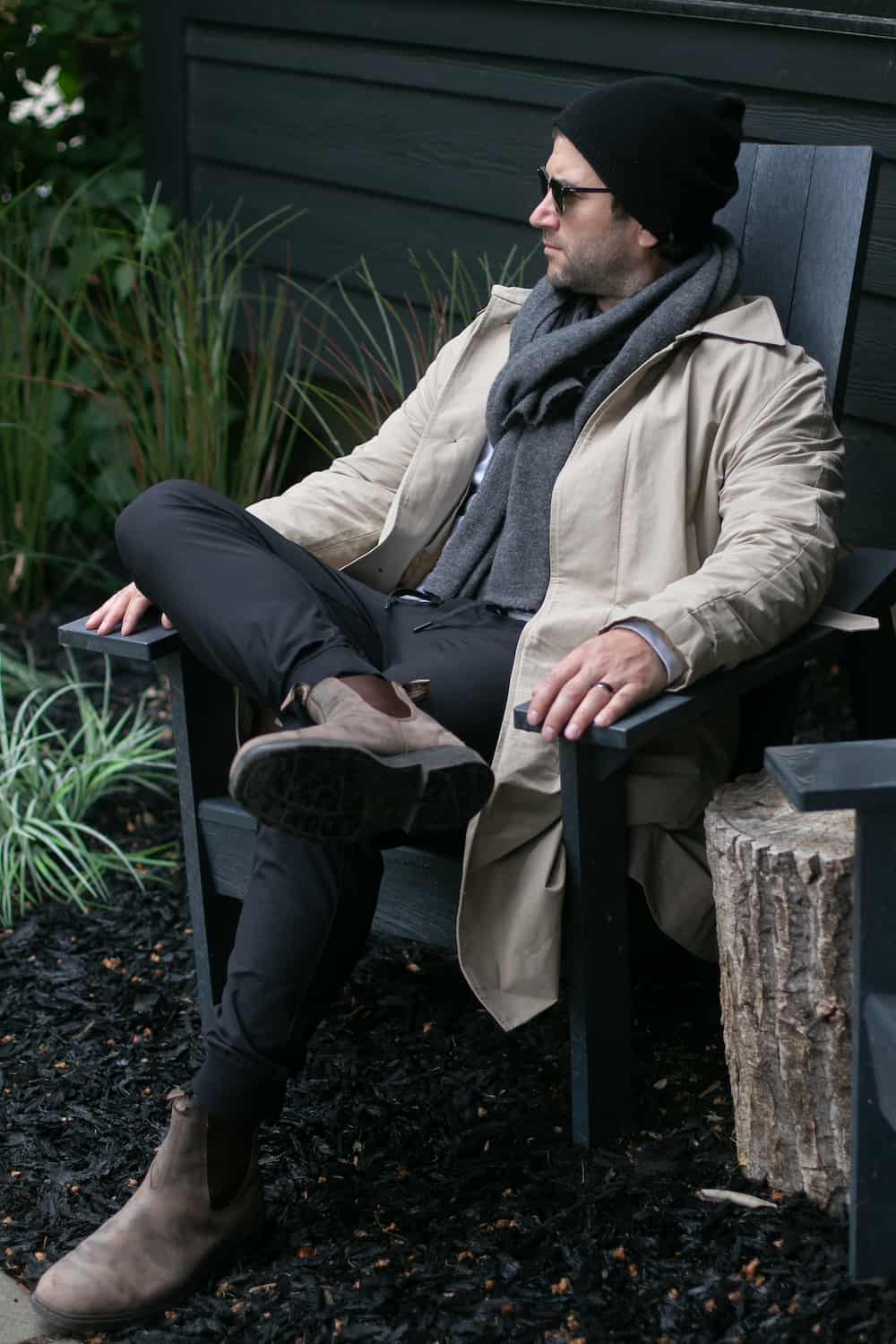 a man wearing black pants, a beige trench coat, light brown Chelsea boots, and a grey scarf with a black beanie