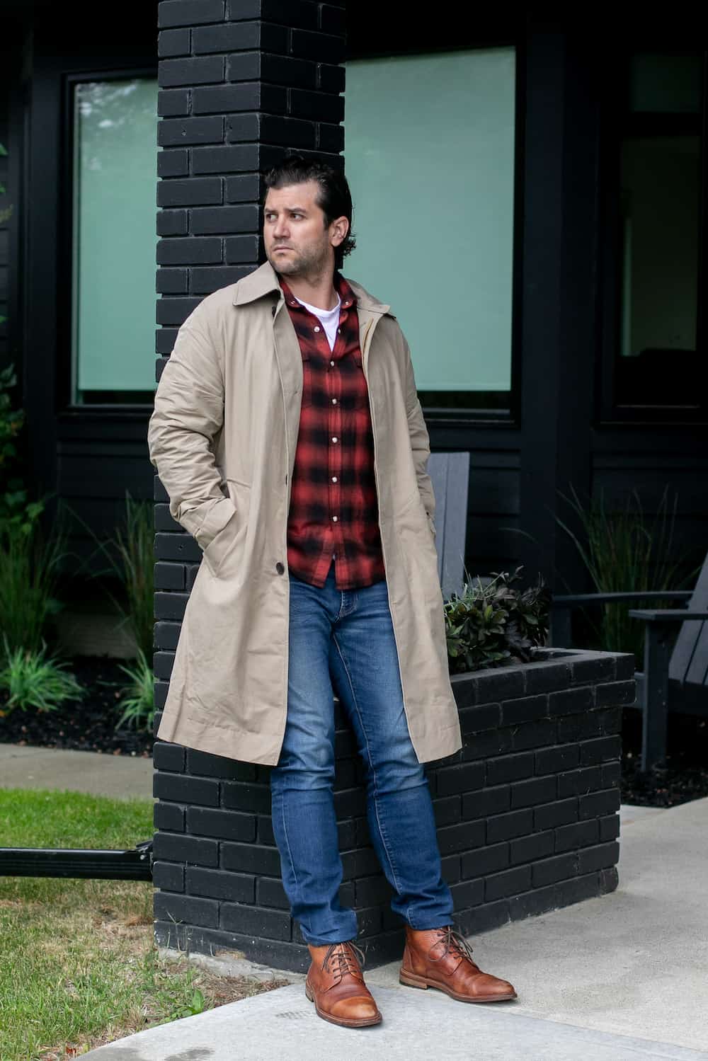 a man wearing blue jeans, red and black flannel, a beige trench coat, and brown boots