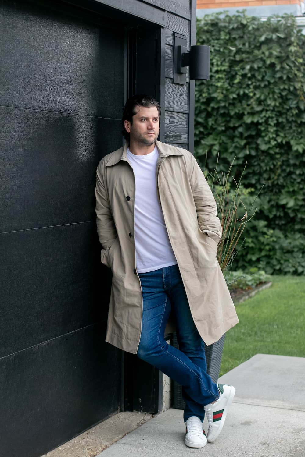 a man wearing blue jeans with a plain white tea, designer sneakers, and a beige trench coat