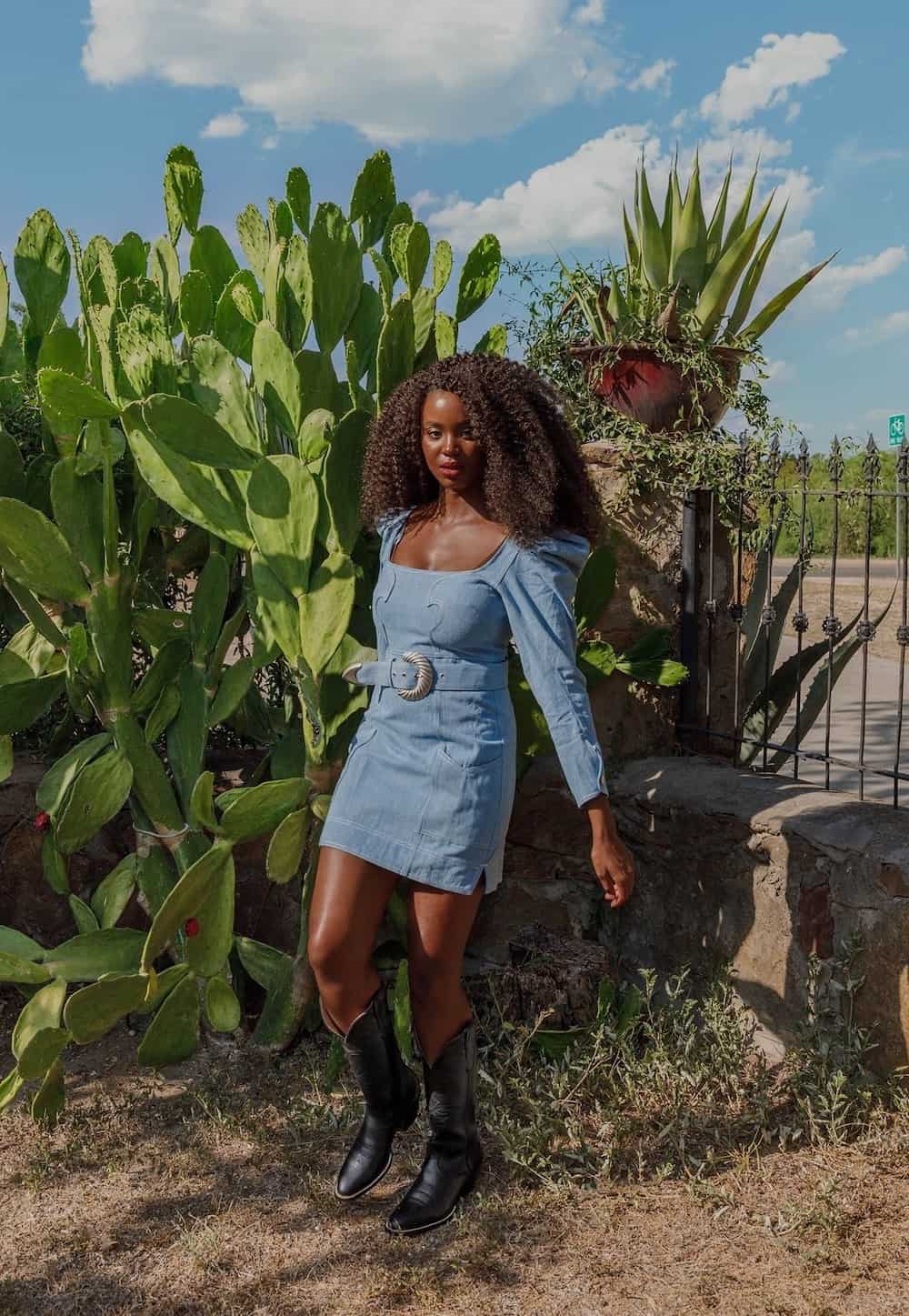 A woman wearing a belted denim mini dress with long puffed sleeves and black cowboy boots