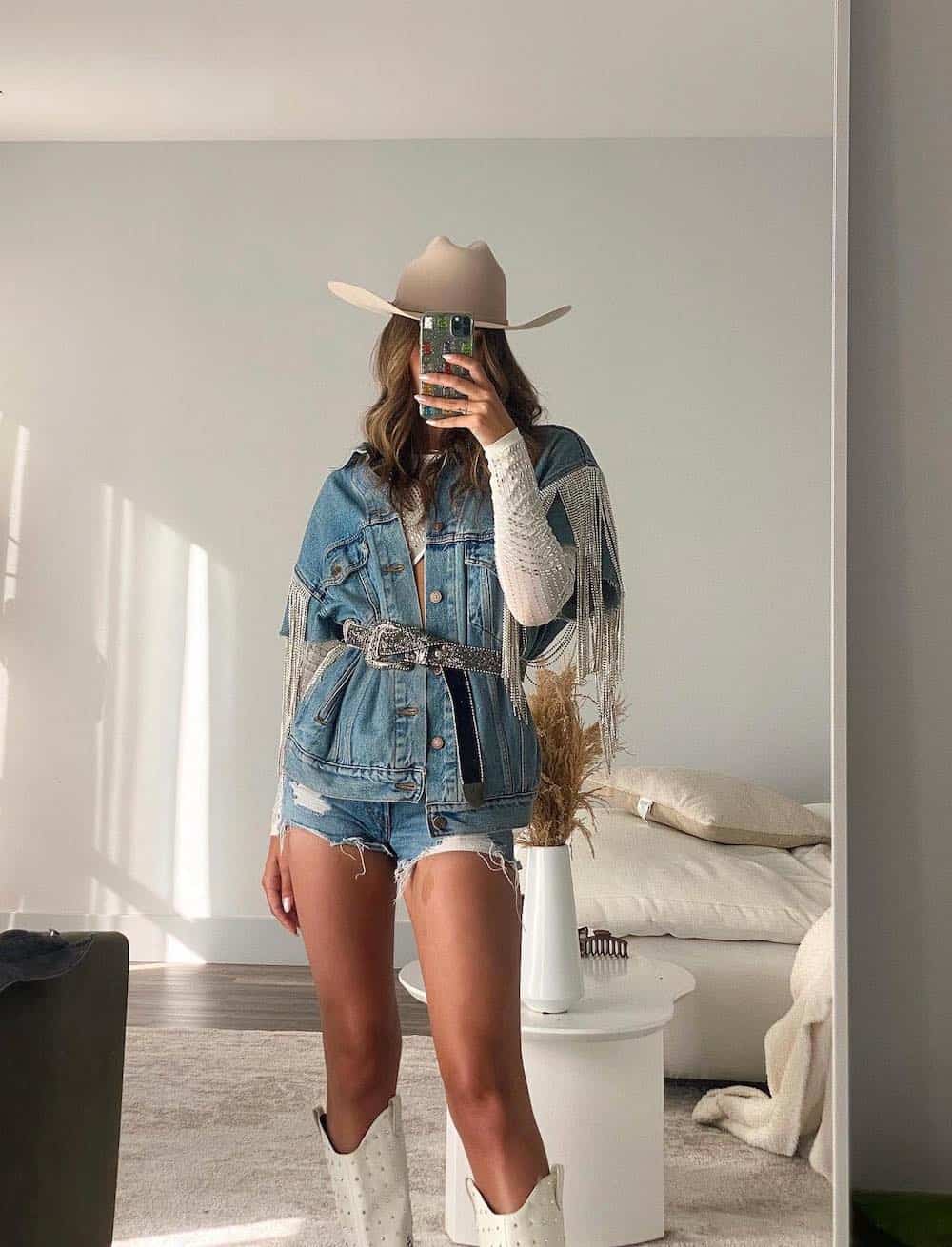 A woman wearing blue denim shorts and a blue denim jacket with silver fringe and a sparkly waist belt with white cowboy boots and a beige cowboy hat