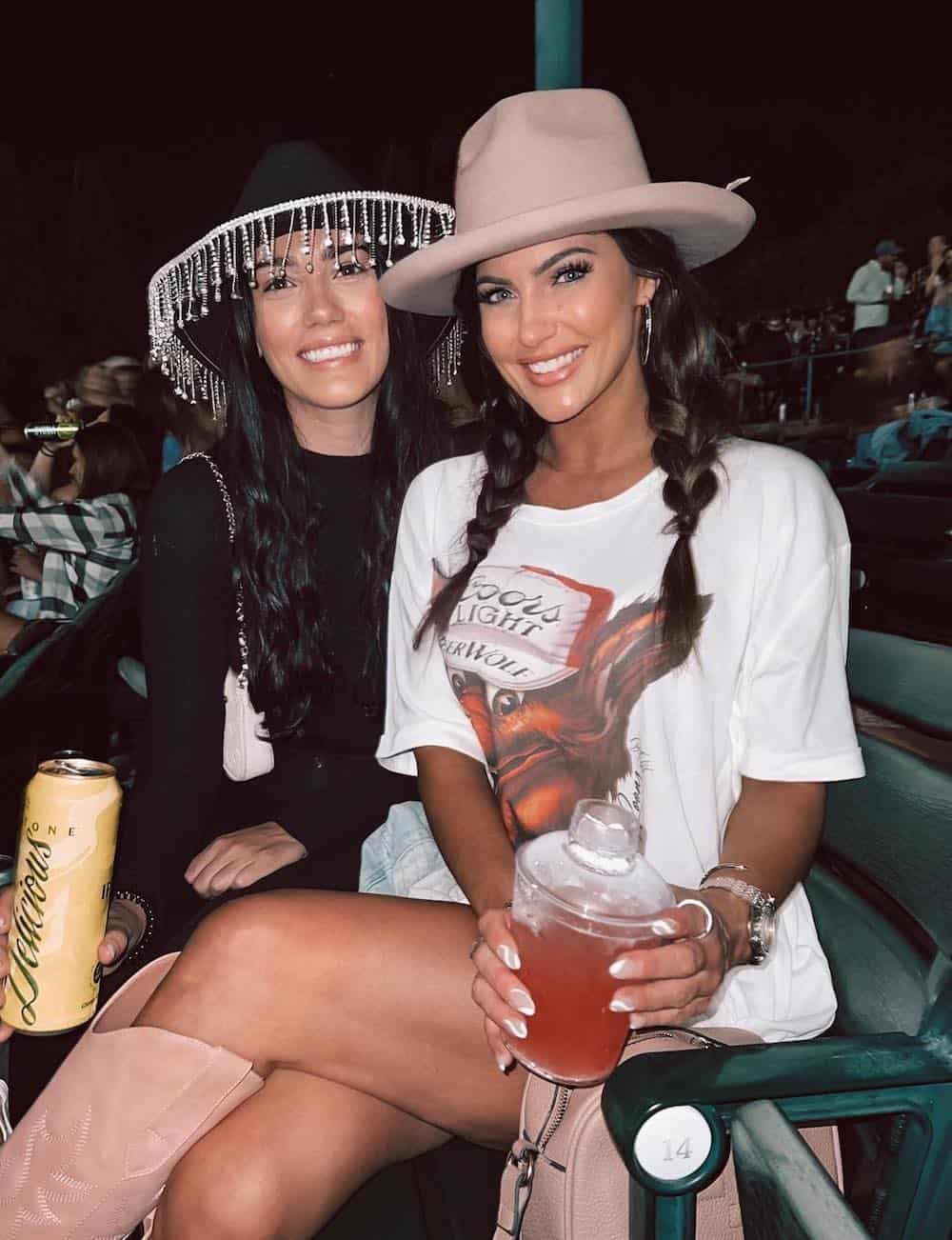 Two women at a concert, one wearing an oversized graphic tee with tall cowboy boots and a fedora and the other wearing a black top with a black cowboy hat featuring gem details