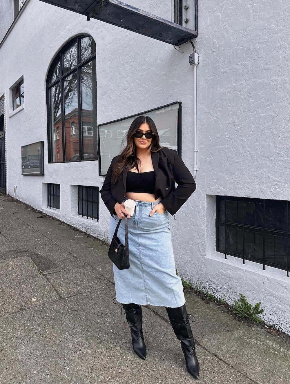 A woman wearing a denim midi skirt with tall black boots, a black bandeau top, a cropped black blazer, and a black shoulder bag