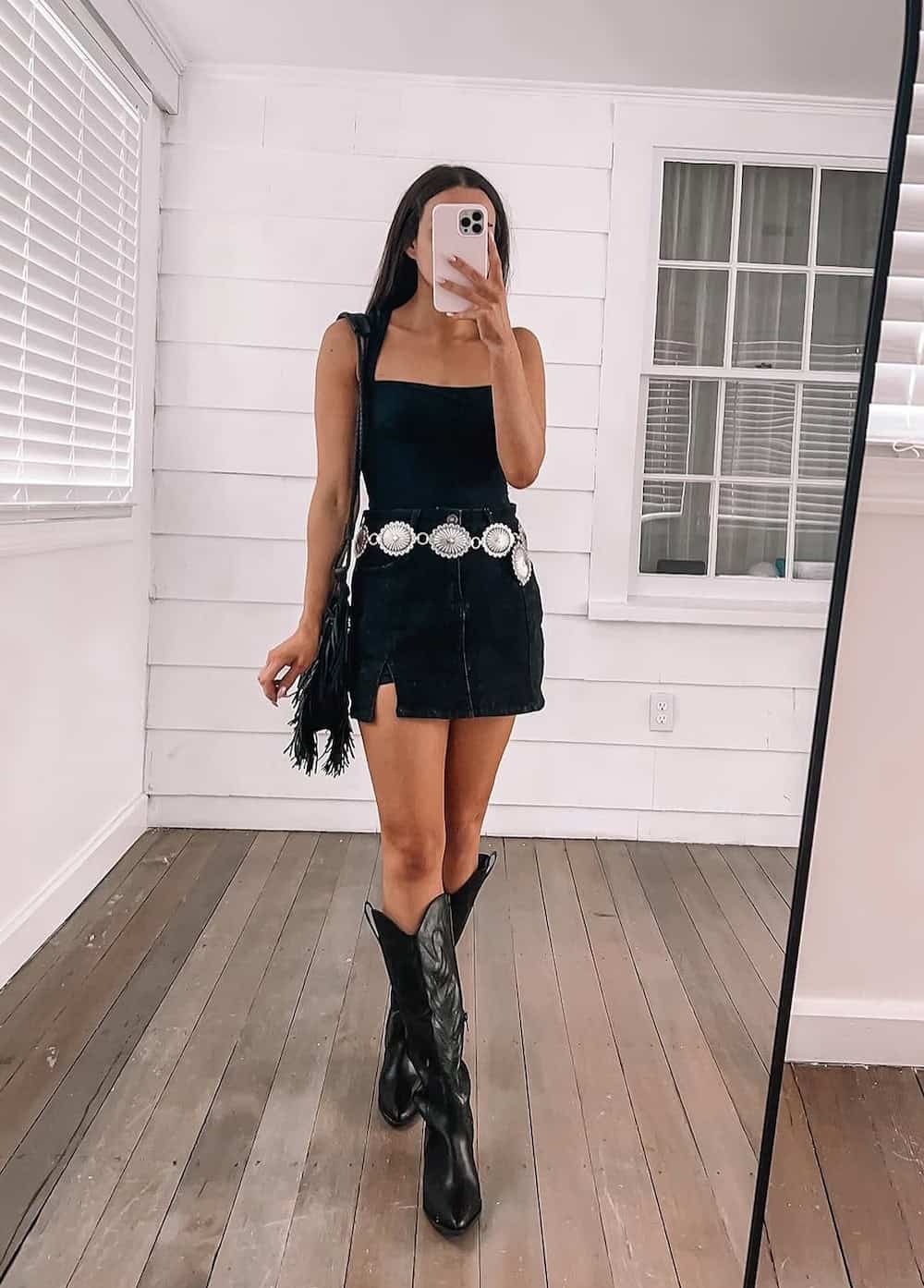 woman wearing a cute country concert outfit with a black tank top and black mini denim skirt with a chain belt and black cowboy boots