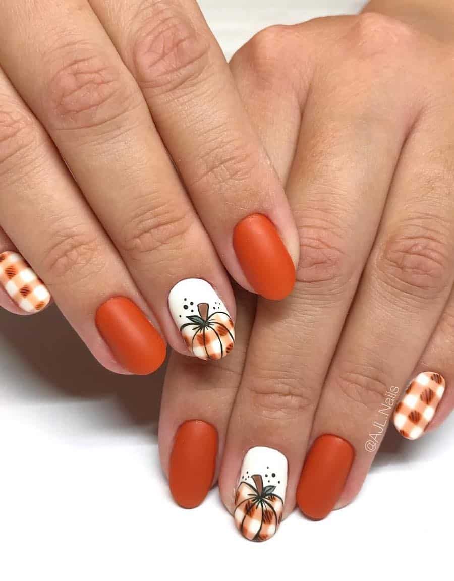 a hand with short round nails painted a matte orange and white with accent nails featuring plaid patterns and plaid pumpkin nail art