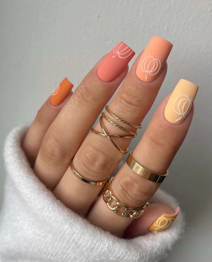 a hand with medium-length square nails painted in a matte gradient design with fall colors with white pumpkin nail art