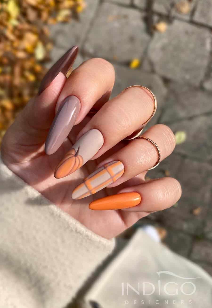 a hand with long almond nails painted with glossy beige, orange, and brown polish with matte beige accent nails painted with pumpkin art and plaid details