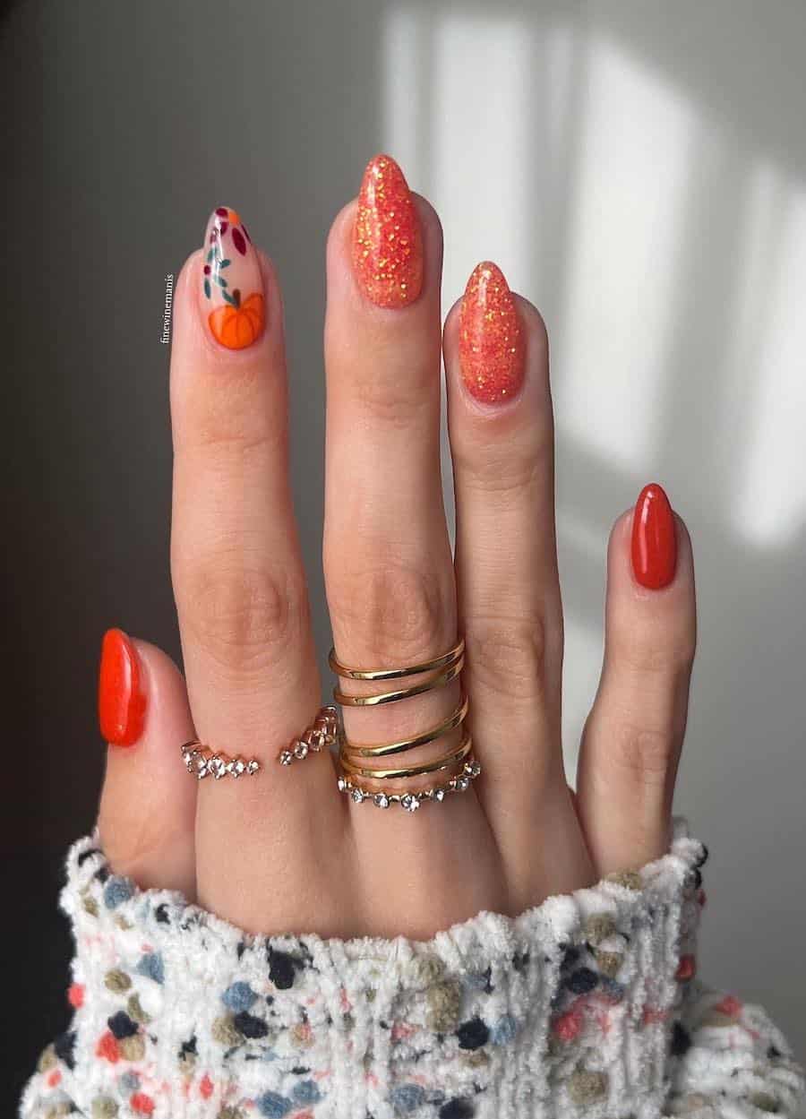 a hand with short almond nails painted solid-colored orange, glittery orange, and a pumpkin art accent nail