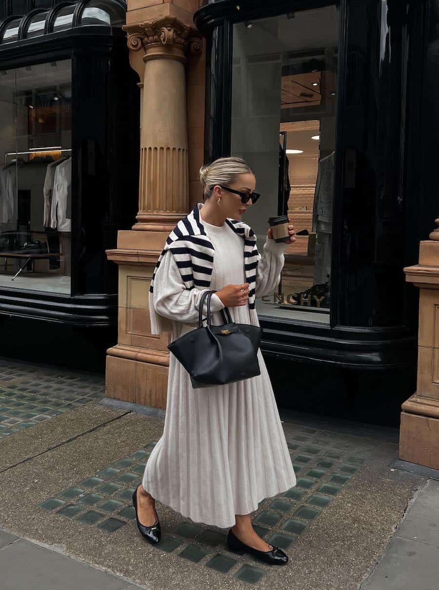 woman wearing a long cream sweater dress with black ballet flats and a black bag