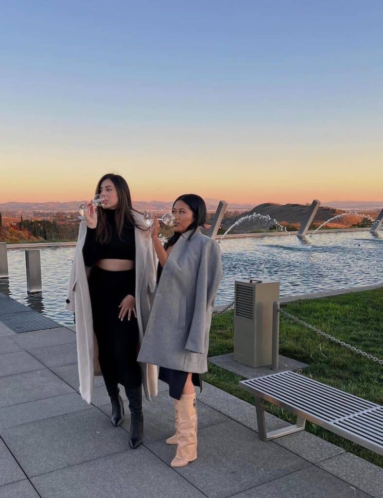 two women at a winery in the fall wearing long coats over sweaters and midi skirts with boots