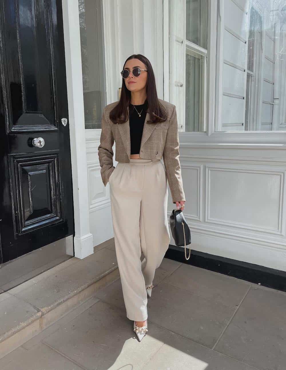 A woman wearing white pants with a black crop top, a beige plaid cropped blaer, white heels, and a black handbag