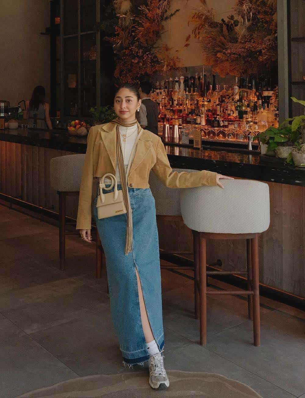 A woman wearing a denim midi skirt, a white top, a brown suede cropped blazer, grey trainers, and a beige crossbody purse