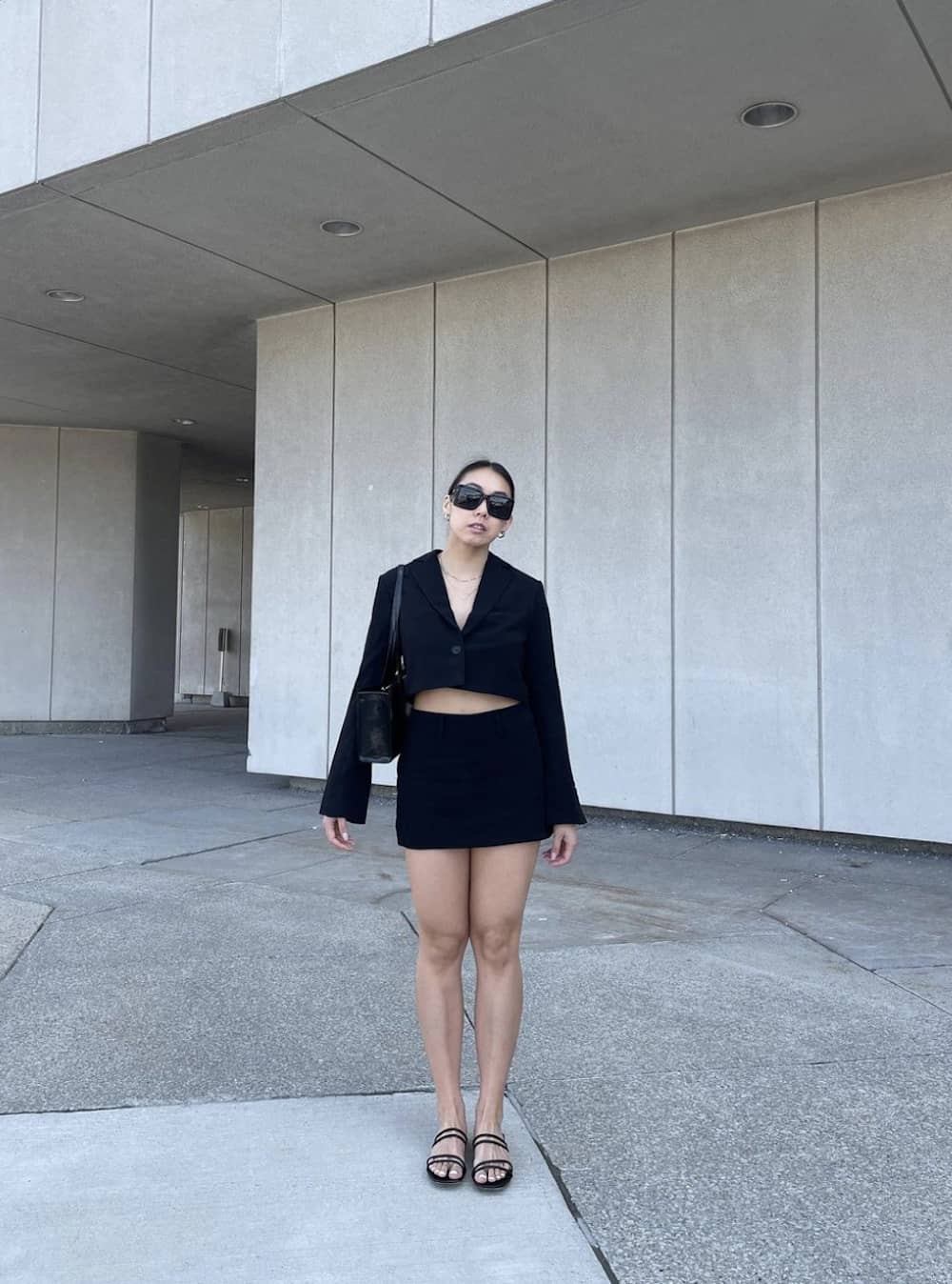 A woman wearing a black mini skirt, a cropped black blazer, black sandals, and a black tote bag