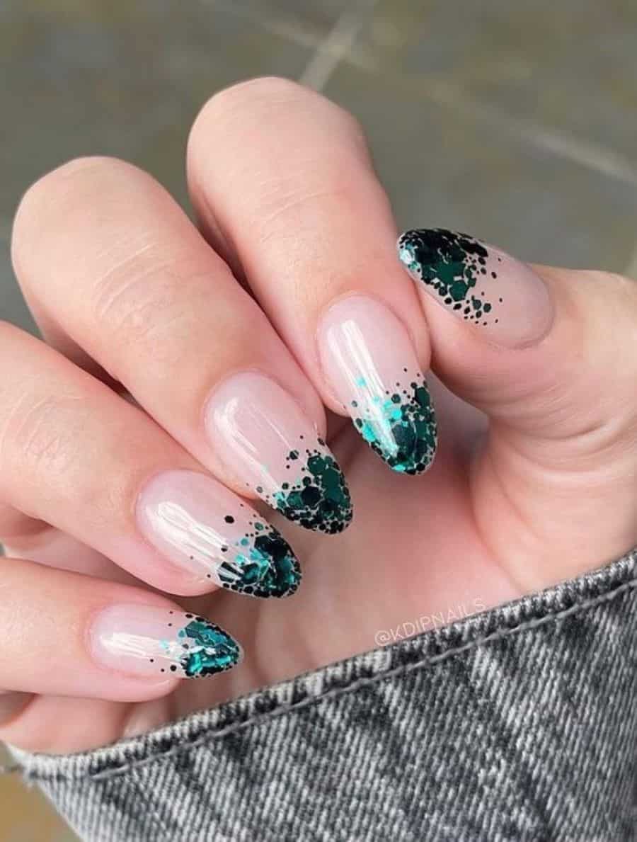 A hand with medium almond nails painted a glossy nude with emerald green chunky glitter French tips