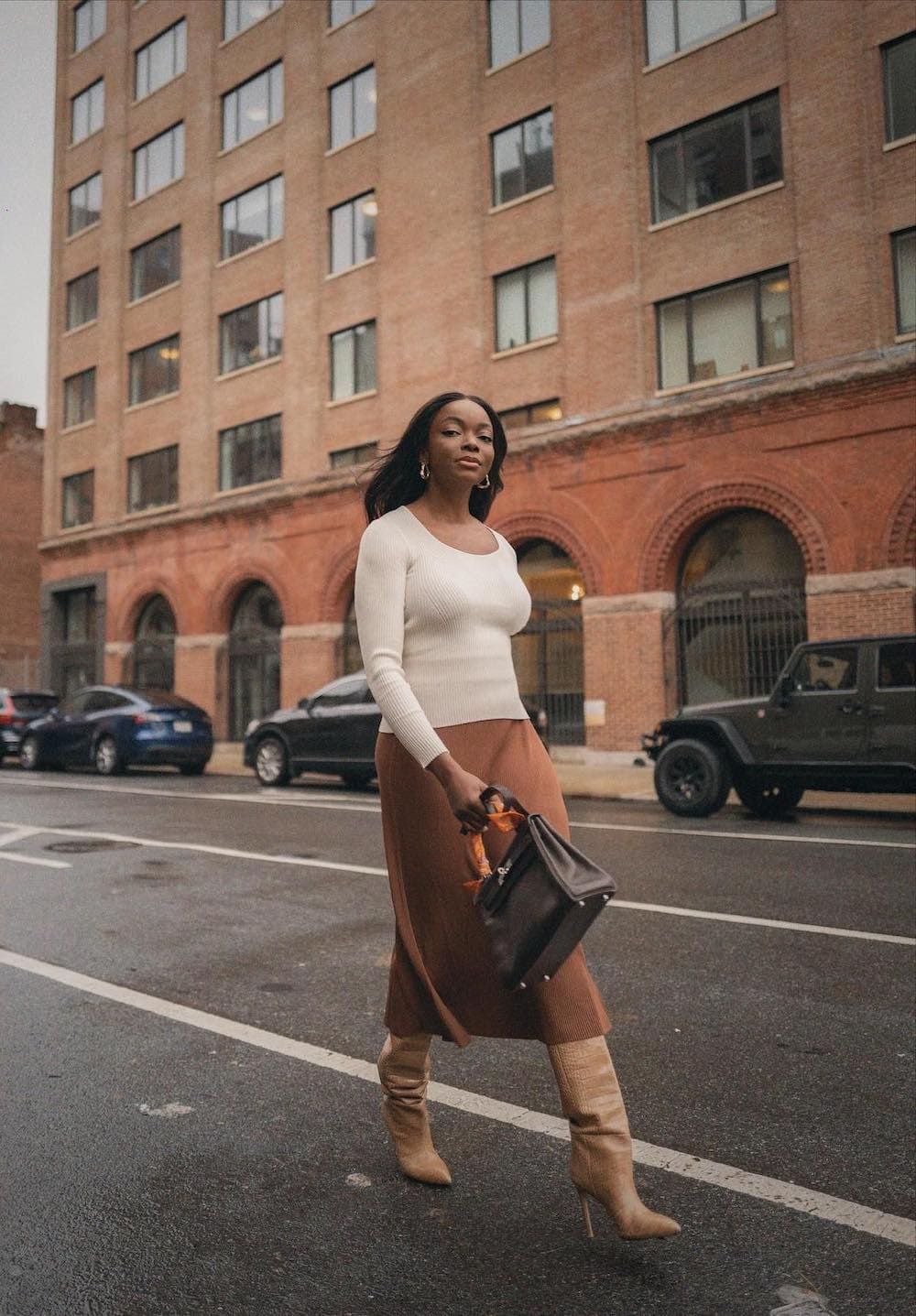 Woman walking down the street wearing a brown midi skirt with tall boots and a white long sleeve sweater.