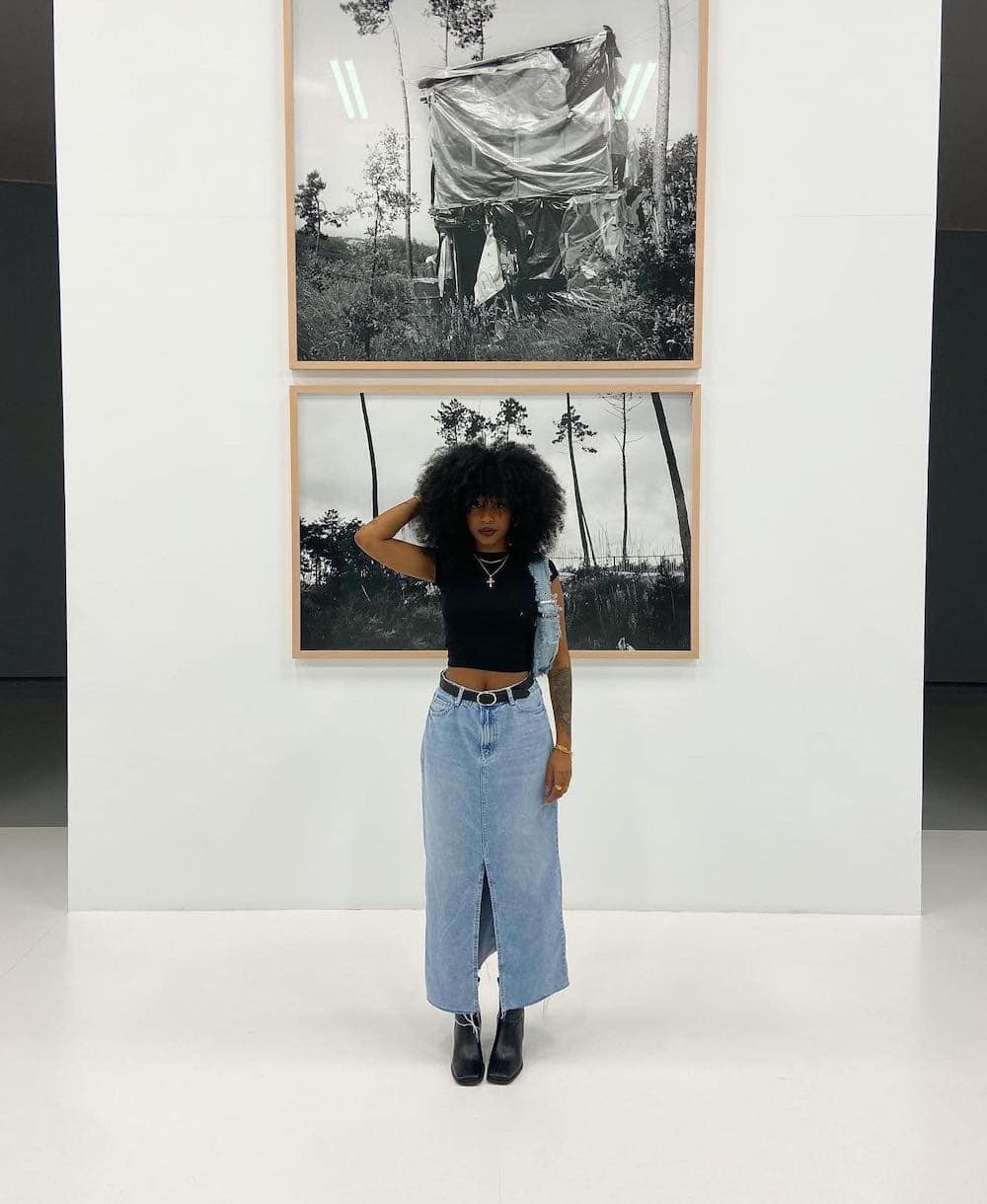 Woman wearing a long denim skirt with a black crop top and black boots in a museum.