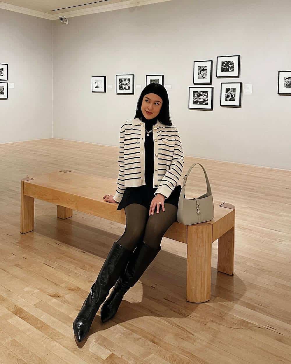 Woman wearing a black dress, tights and tall boots with a white and black and striped sweater in a museum.