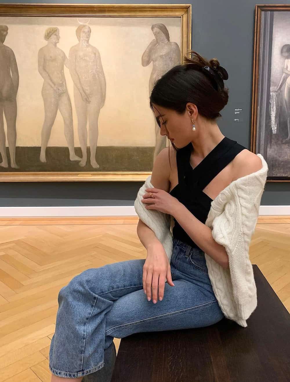 Woman wearing straight leg jeans with a black tank top and a white sweater in a museum.