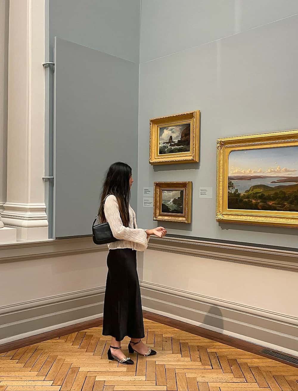 Woman wearing a black silk slip dress and a cream sweater with Mary Jane shoes in a museum.