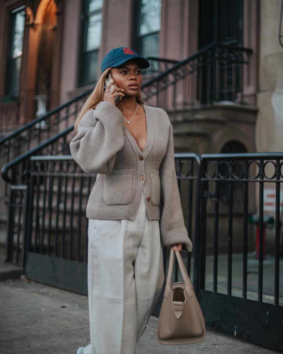 A woman wearing off-white pleated pants with a beige cardigan, a blue baseball cap, and a tan leather tote