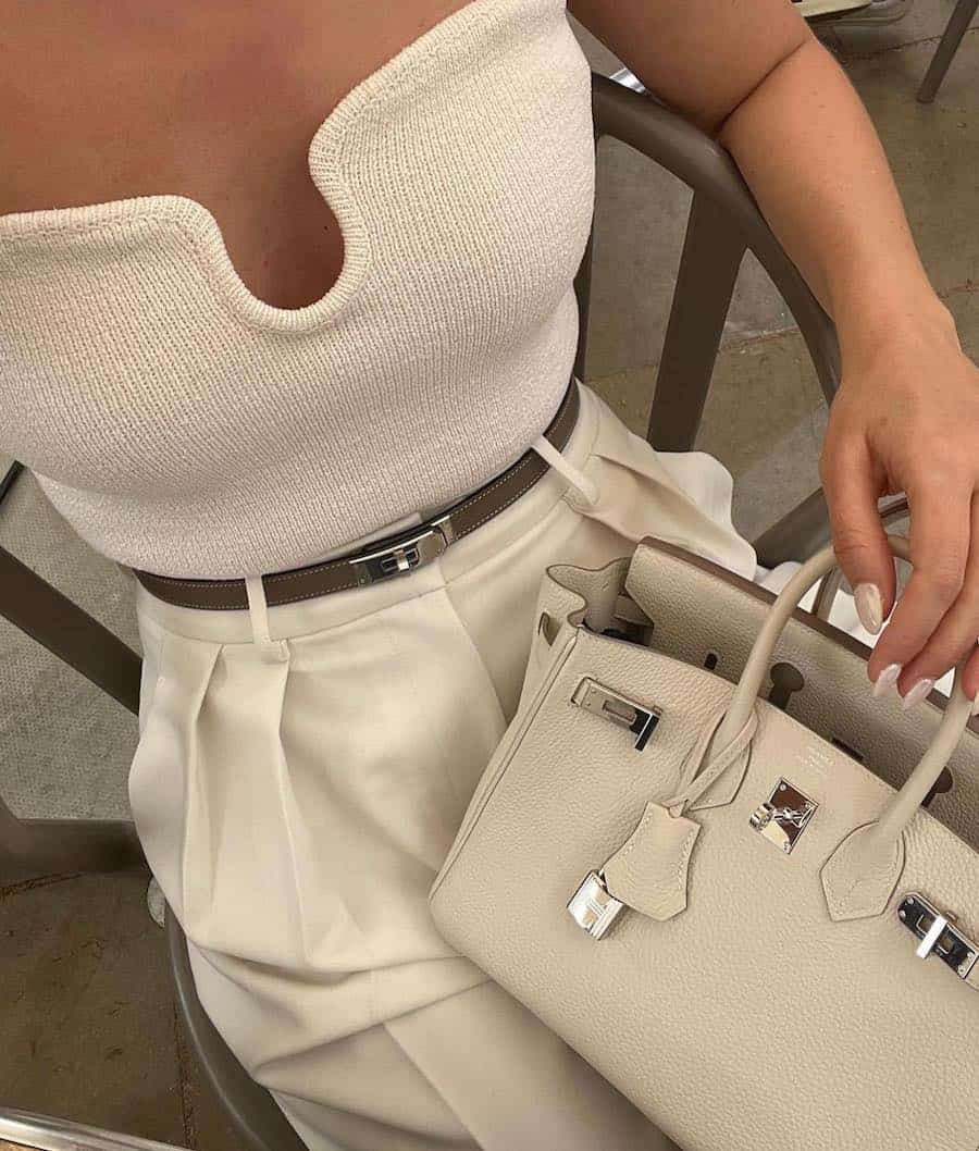 An overhead shot of a woman wearing pleated white pants with a knit top and a white leather Birkin bag