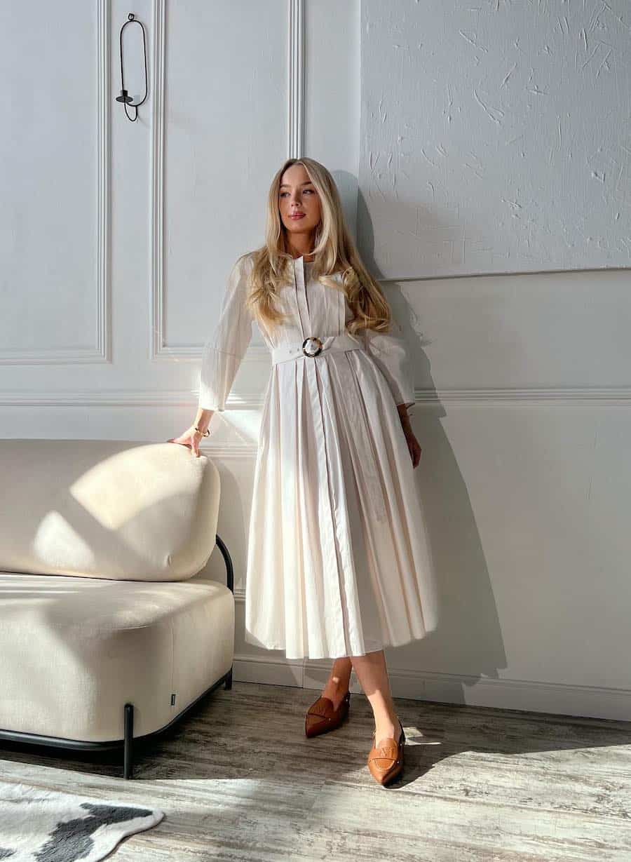 A woman wearing a long-sleeved pleated white dress with a waist belt and brown leather loafers