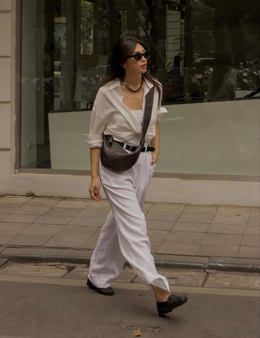 A woman wearing white trousers with an off-white button-up, black loafers, and a brown crossbody bag