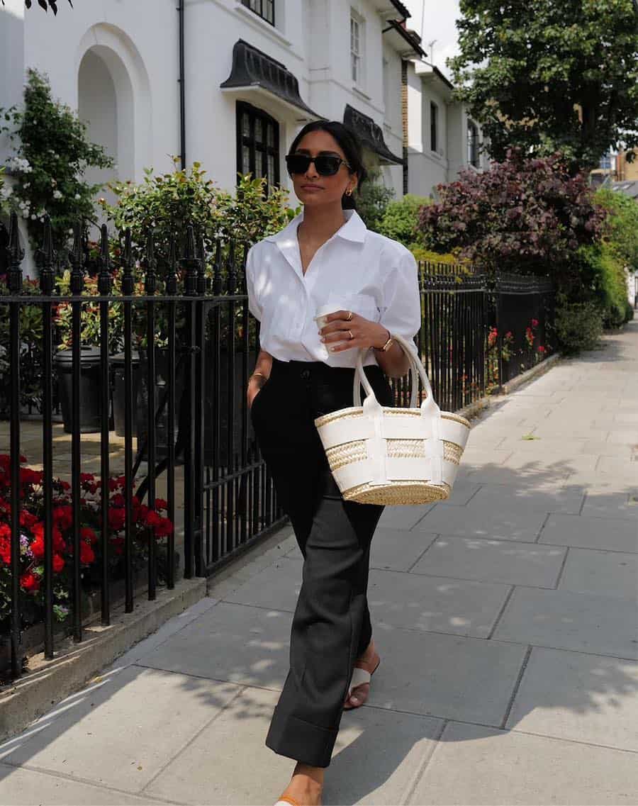 A woman wearing black trousers, a white button-up, and white sandals with a raffia and white leather tote