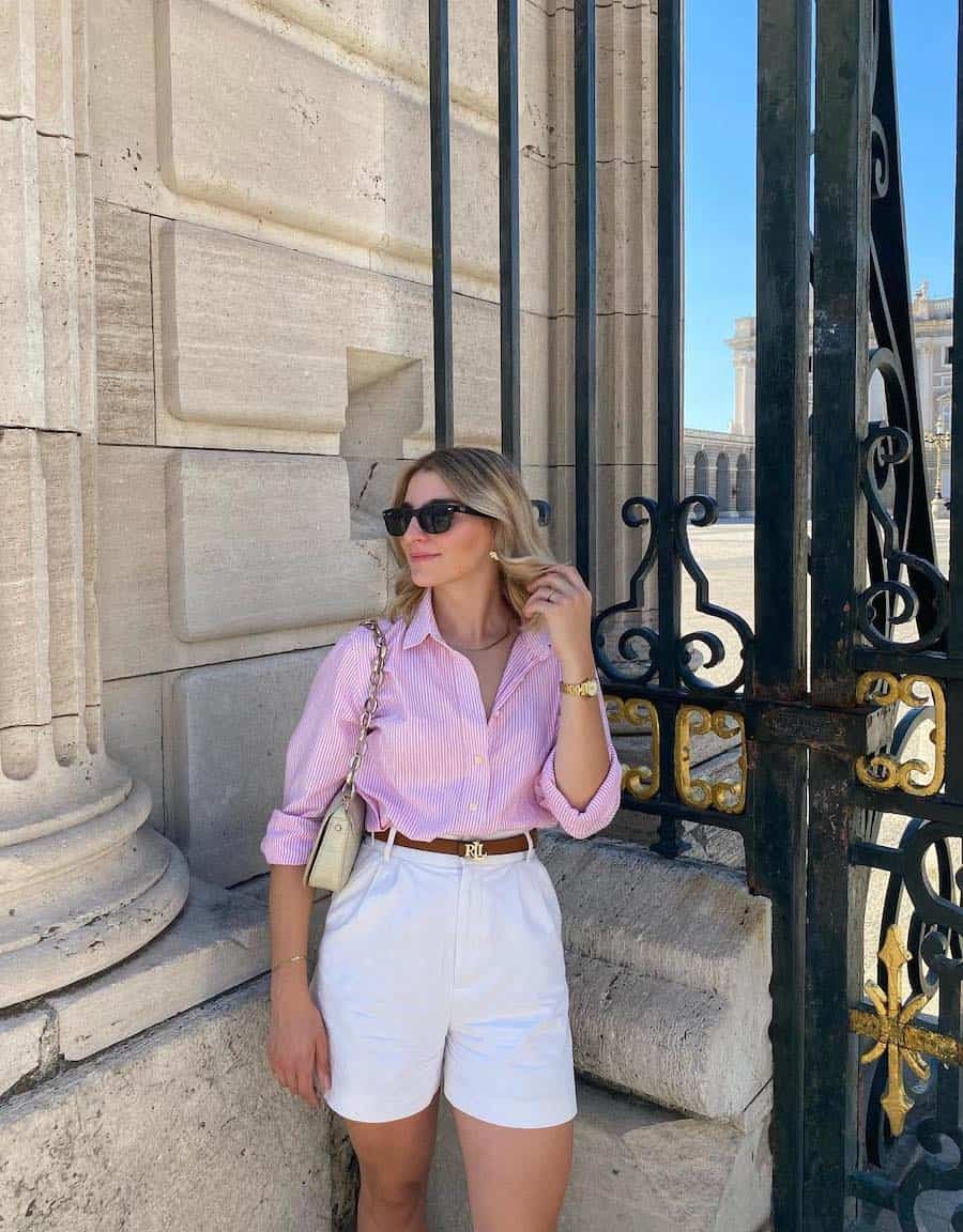 A woman wearing white pleated shorts with a pink striped button-up and a white leather shoulder bag