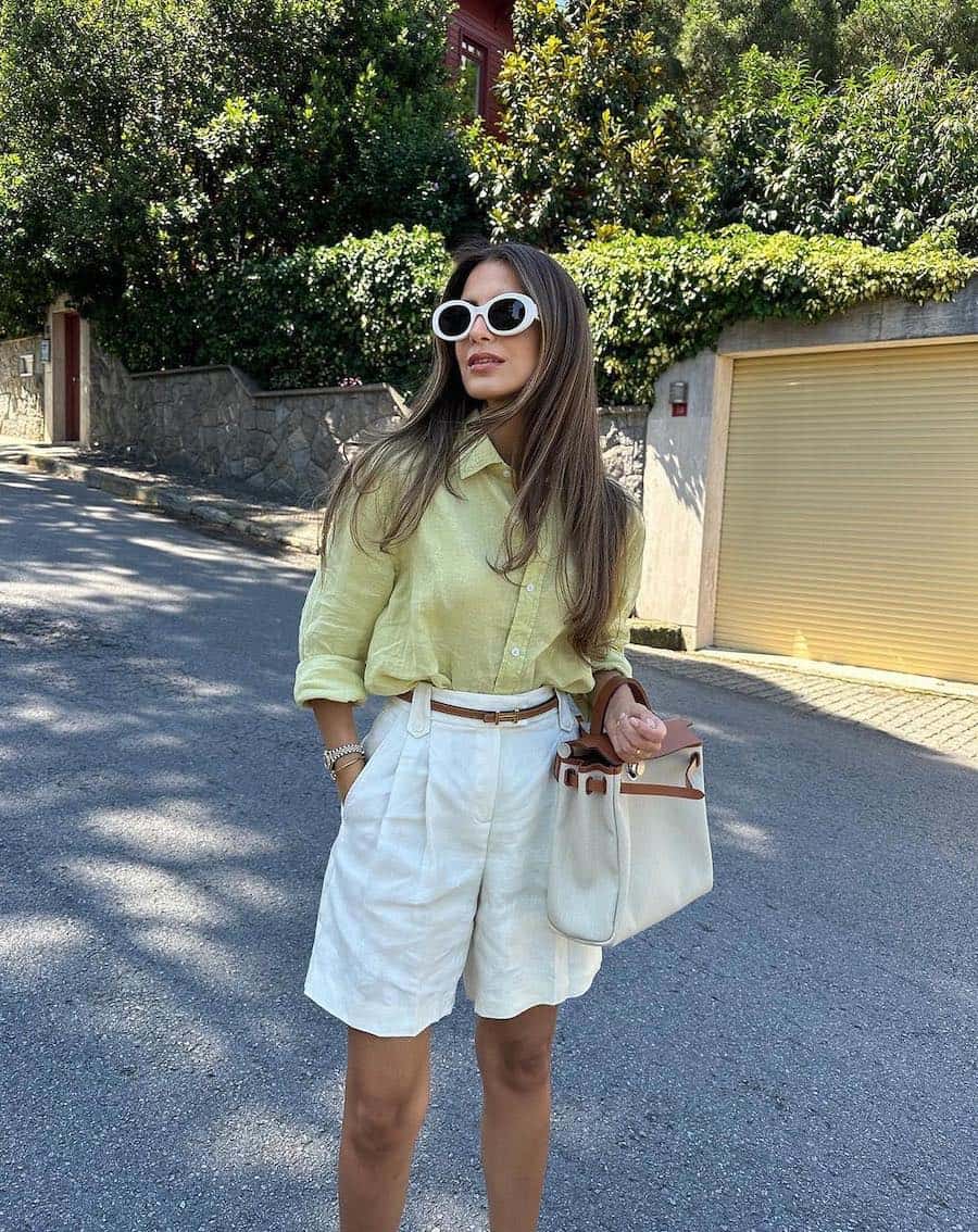 A woman wearing white pleated shorts with a yellow button-up with a white and brown leather tote