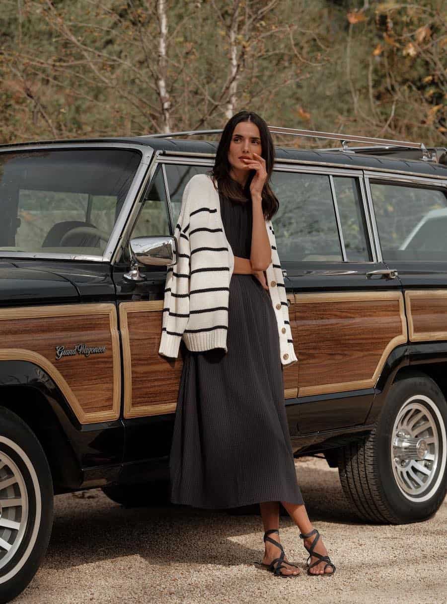 A woman wearing a black midi dress with a black and white striped cardigan and strappy black heels