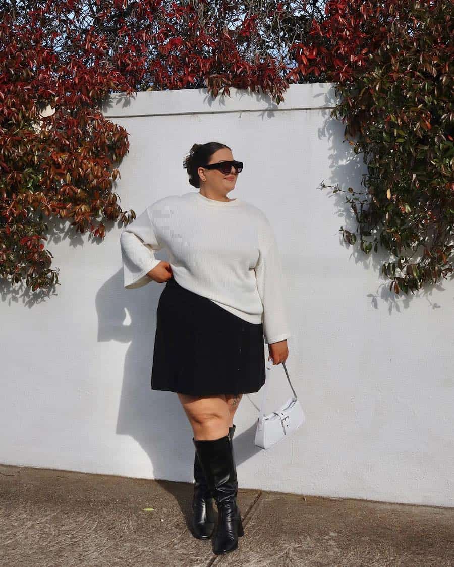 A woman wearing a black mini skirt, a white sweater, and tall black boots with a white handbag