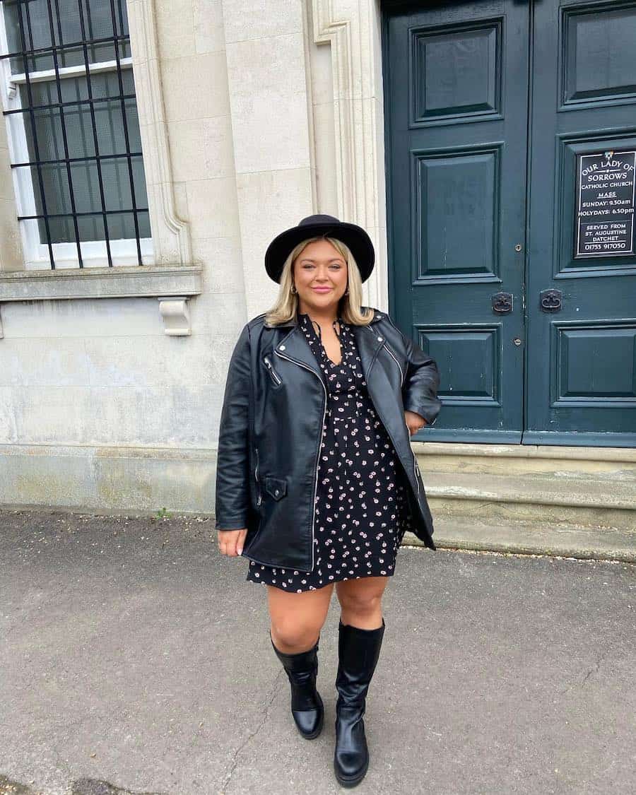 A woman wearing a black floral mini dress with tall black leather boots, a black leather jacket, and a black fedora