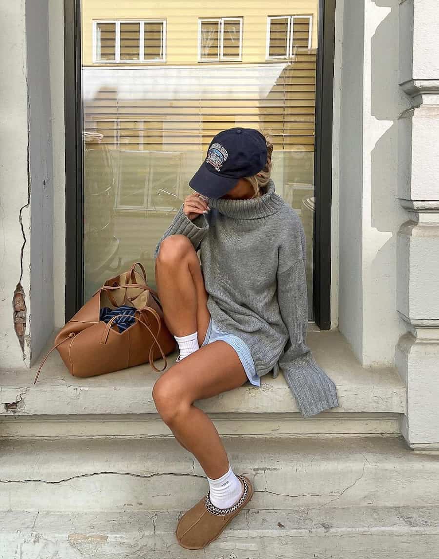 A woman wearing blue linen shorts with an oversized grey turtleneck sweater, a baseball cap, and brown slippers
