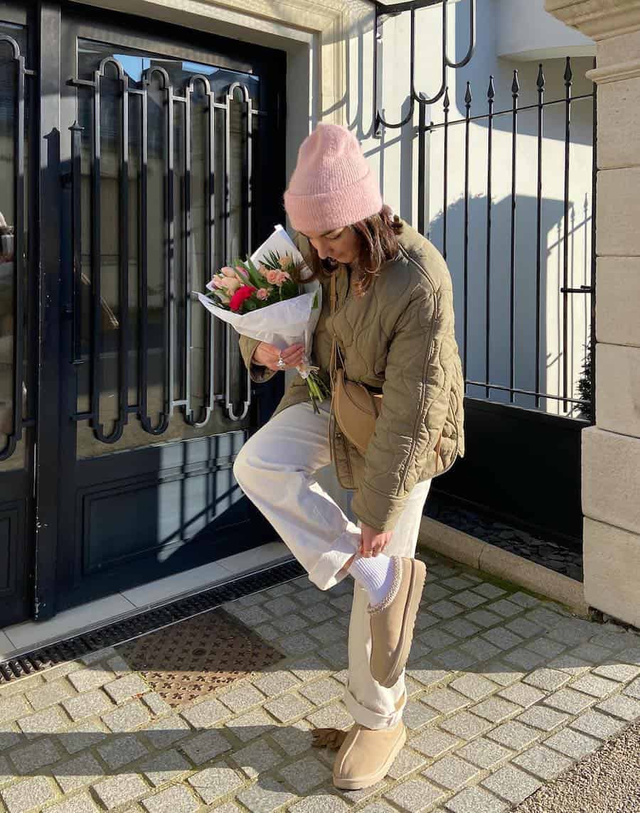 A woman wearing white jeans with brown Ugg Tasman slippers, a quilted brown jacket, and a pink beanie