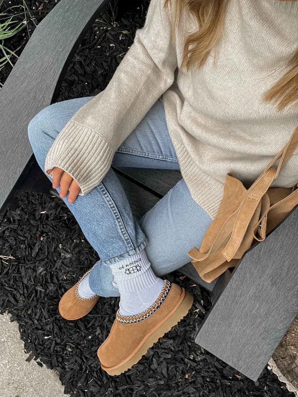 An overhead shot of a woman wearing brown Ugg Tasman slippers with blue jeans and a fine knit beige sweater