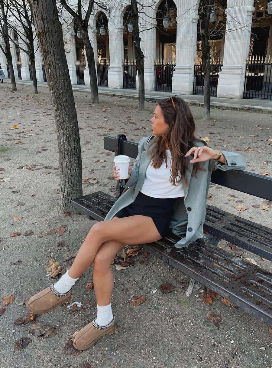 A woman wearing black shorts, a white crop top, a light blue blazer, and brown Ugg Tasman slippers