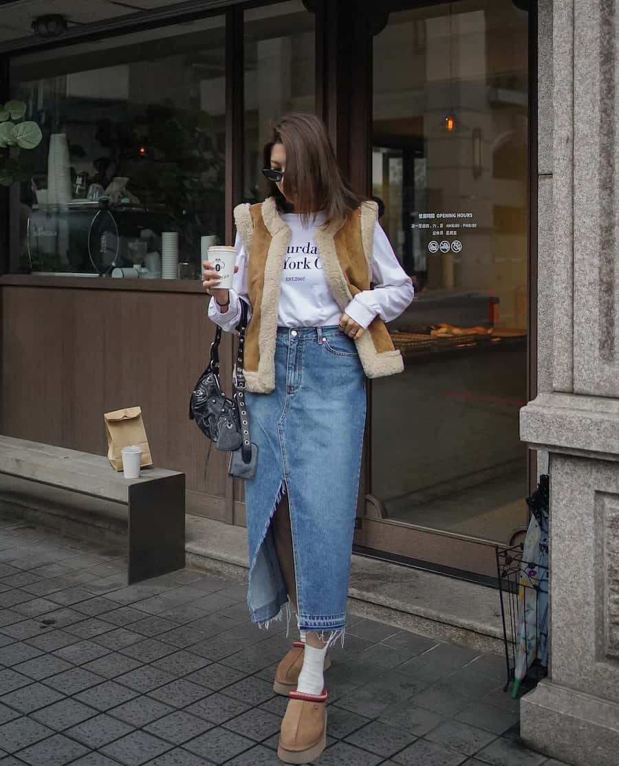 A woman wearing a denim midi skirt with a long sleeve graphic tee, a brown vest with sherpa, and brown Ugg Tasman slippers