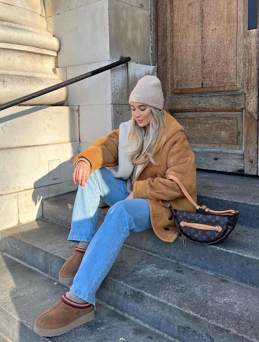 A woman wearing light blue jeans with a brown sherpa lined coat, a beige beanie, and brown Ugg Tasman slippers