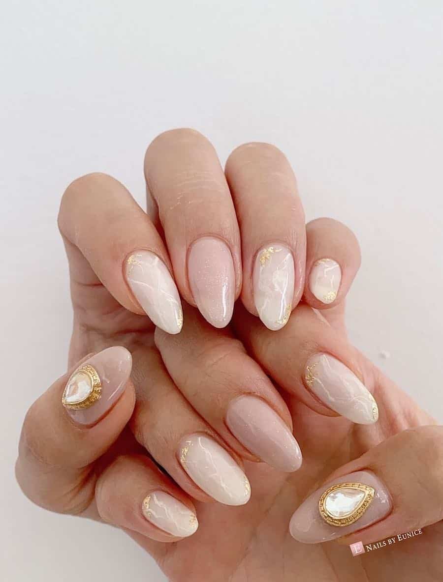 Medium length almond nails with white marbling, nude accent nails, gold flake details, and gold and clear gem accents