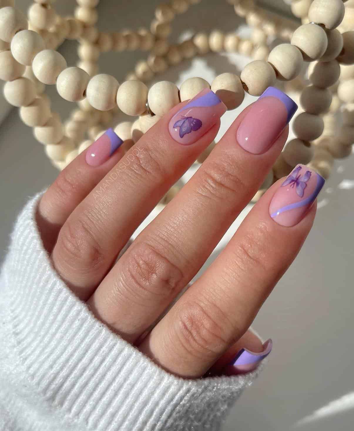 A hand with short square nails painted a nude pink polish with light purple French tips and waves with purple butterflies