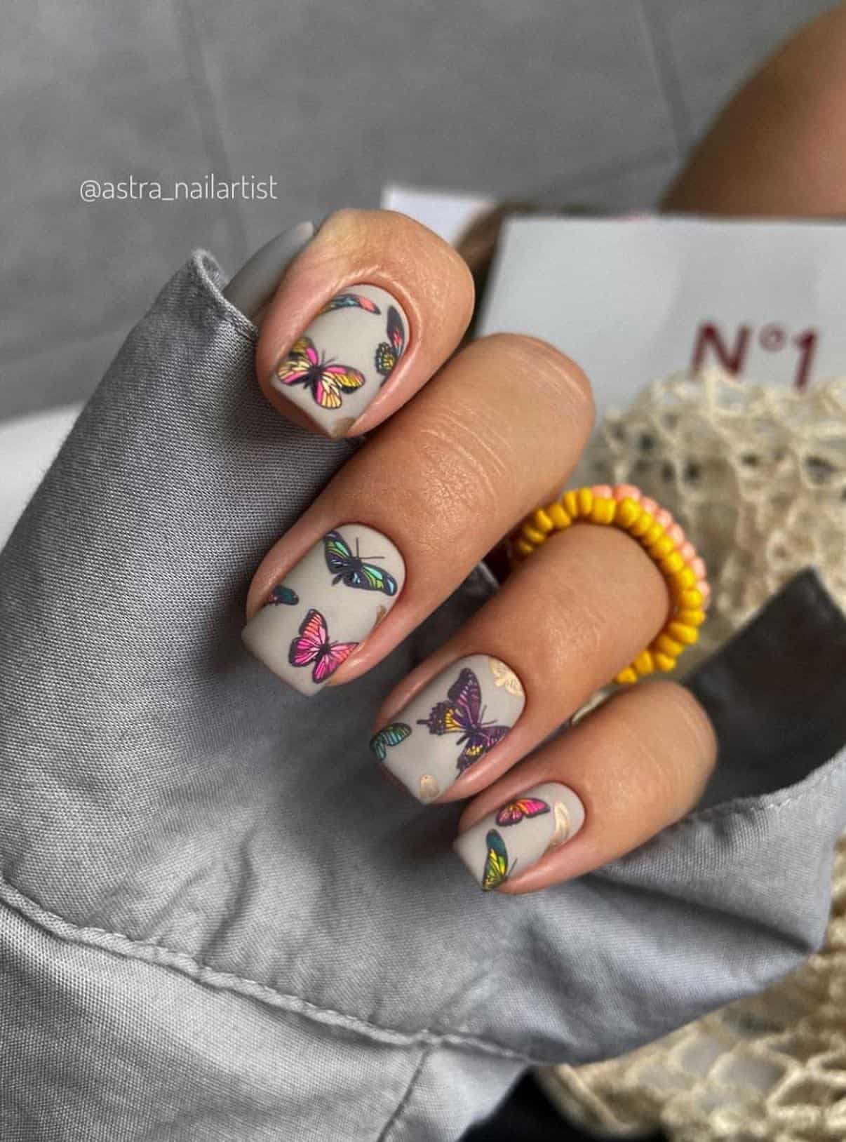 A hand with short matte grey nails with colorful butterflies