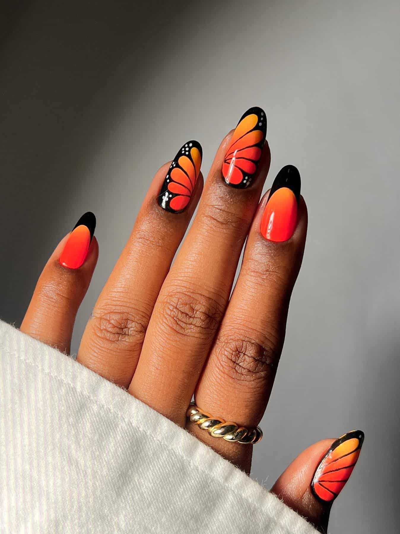 A hand with medium length almond nails painted a red and orange ombre with black French tips and black butterfly wing art accent nails