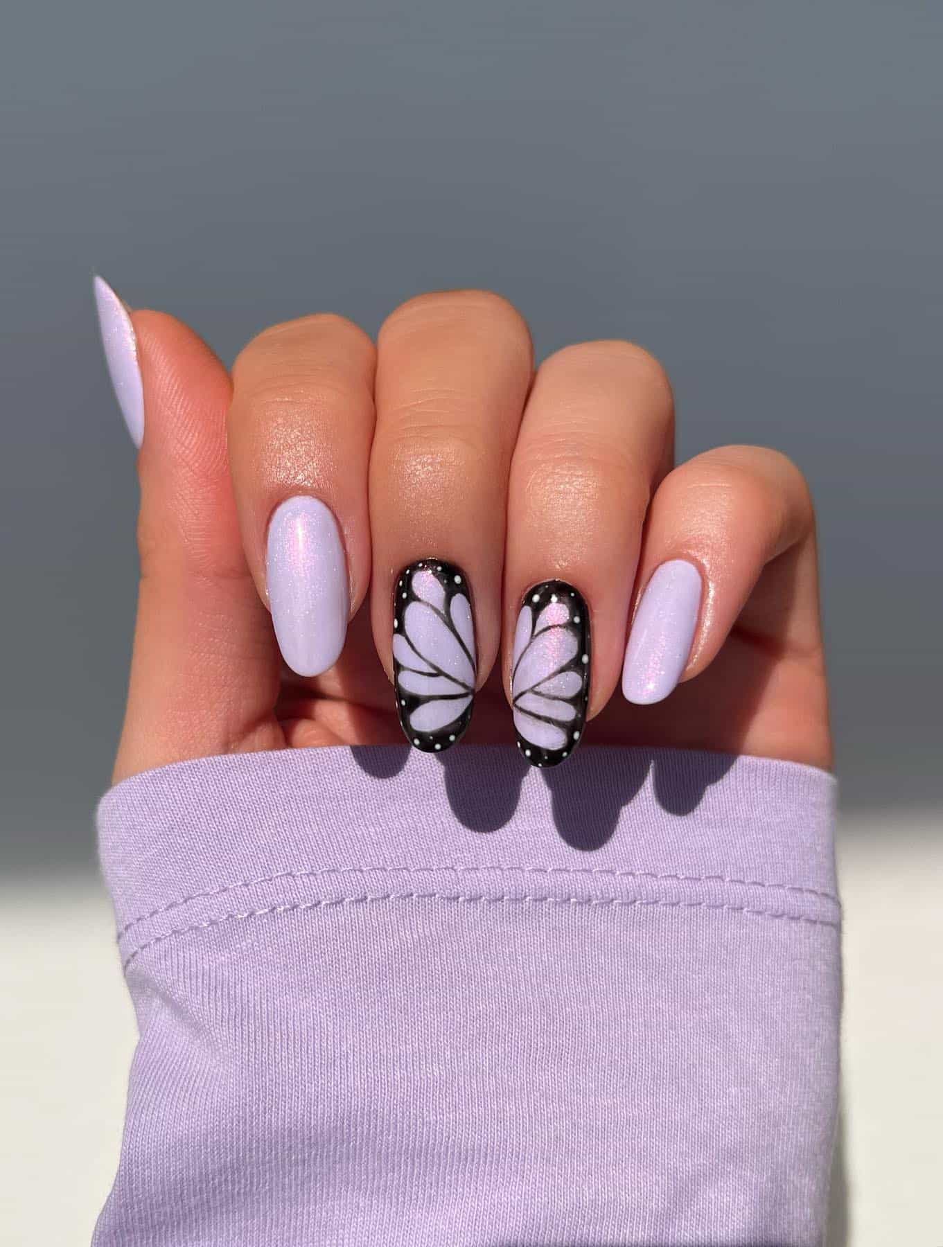 A hand with medium length almond nails painted a shimmering light purple with butterfly wing accent nails
