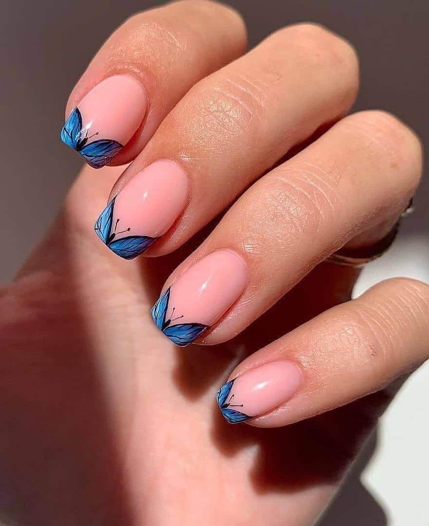 A hand with short square nails painted a nude pink with blue butterfly French tips