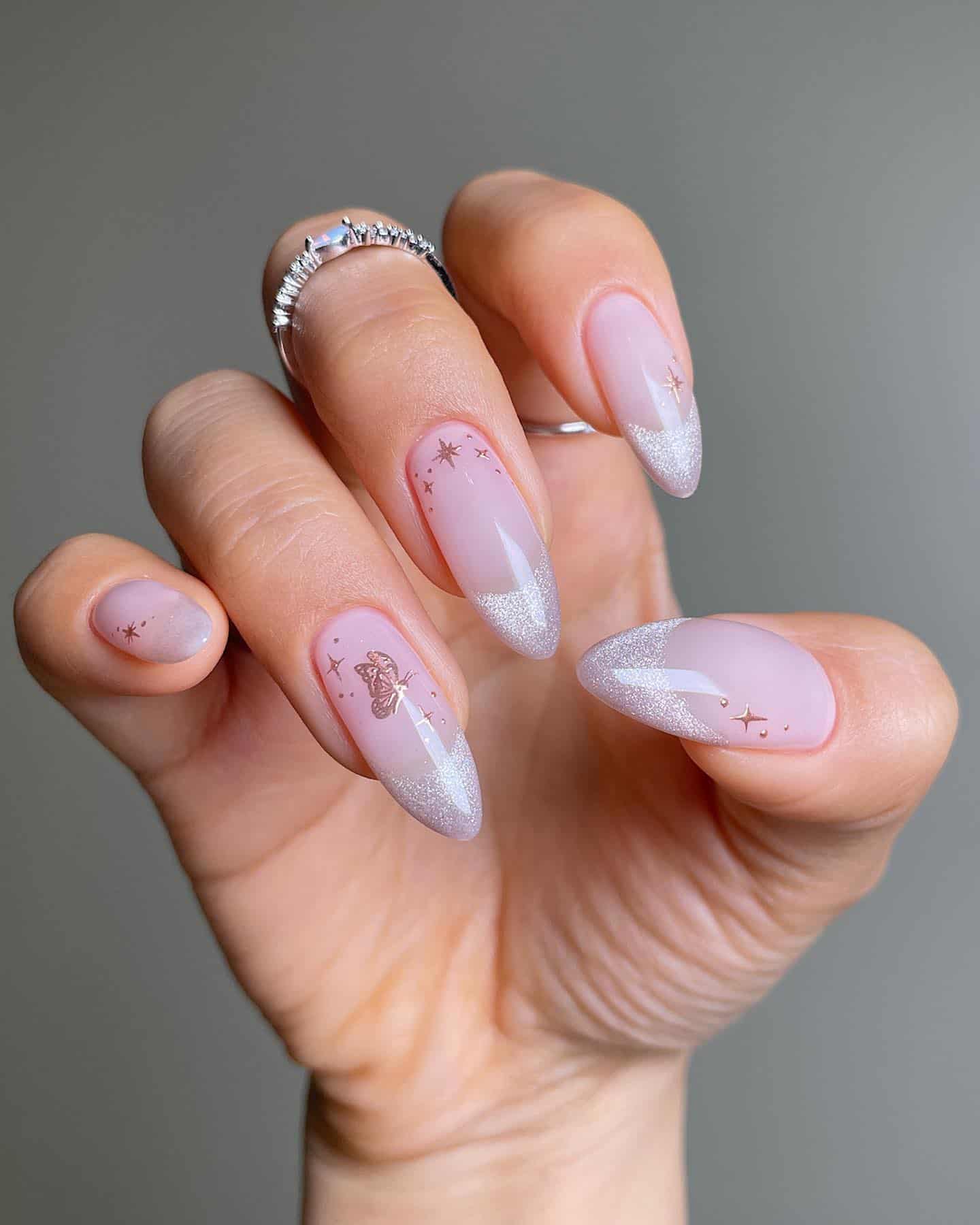 A hand with long nude pink almond nails with shimmering pearl French tips and bronze sparkle and butterfly accents
