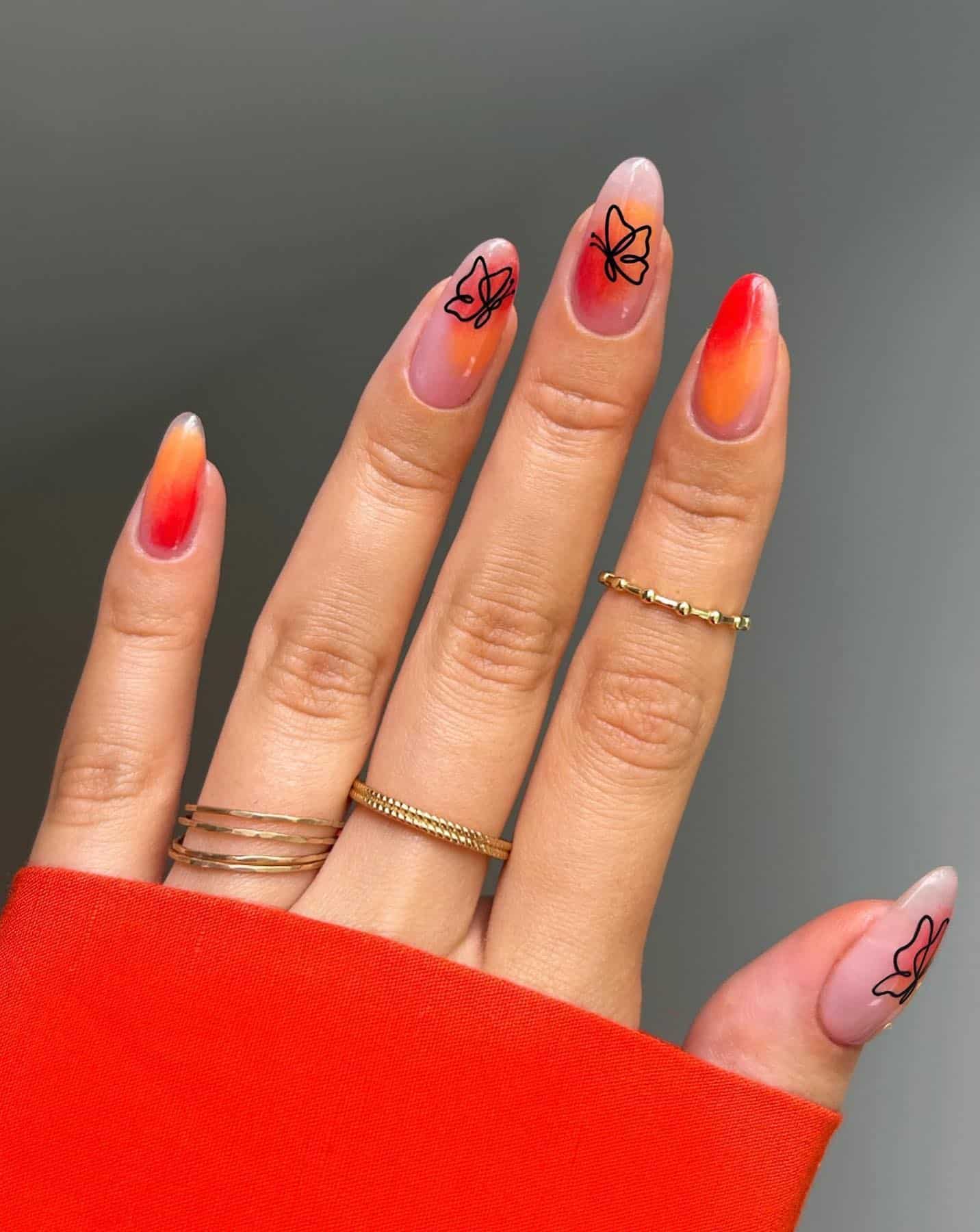 A hand with short almond nails painted a nude polish with red and orange abstract details and black butterfly outlines
