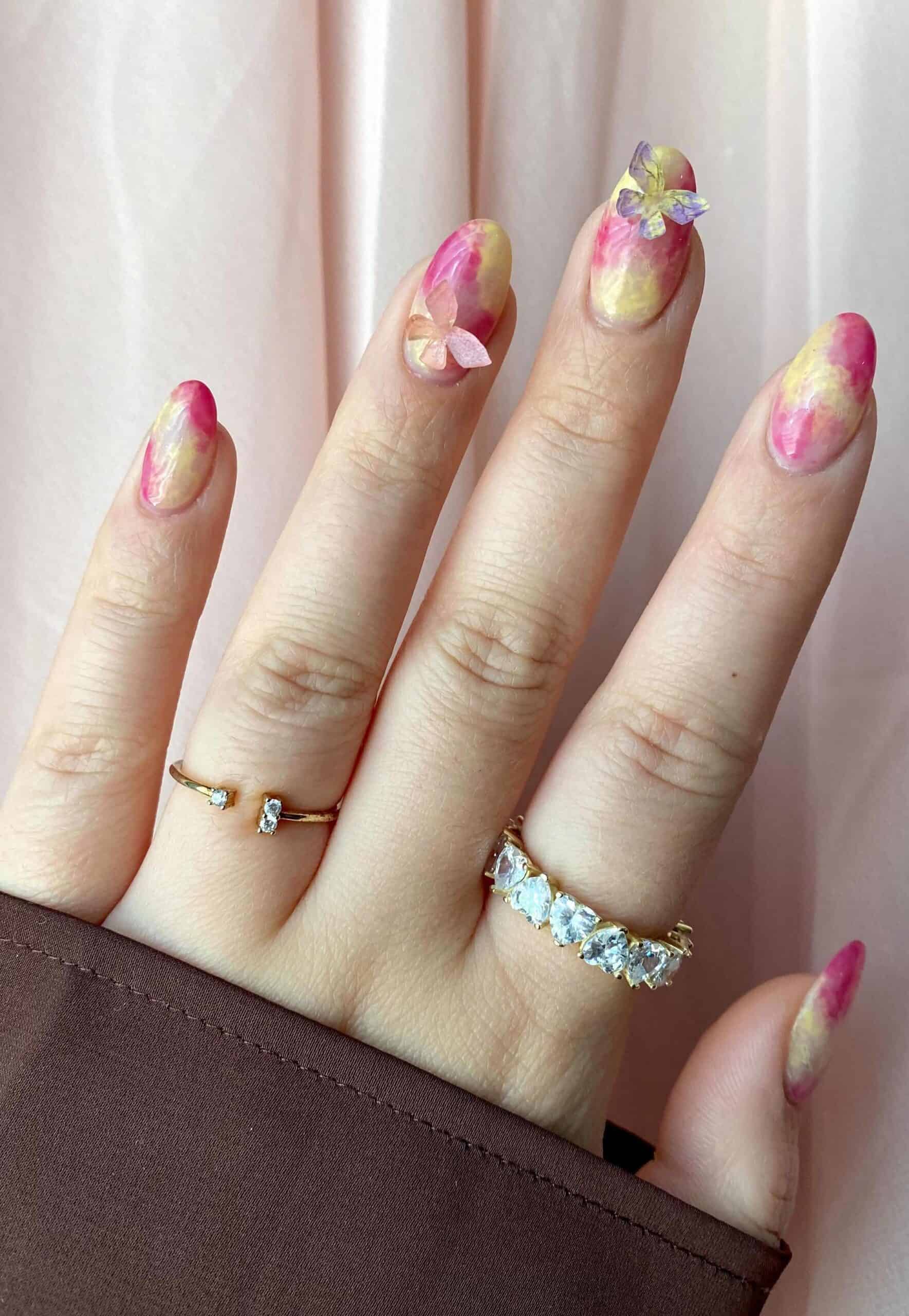A hand with short round yellow and pink marbled nails with 3D butterfly accents