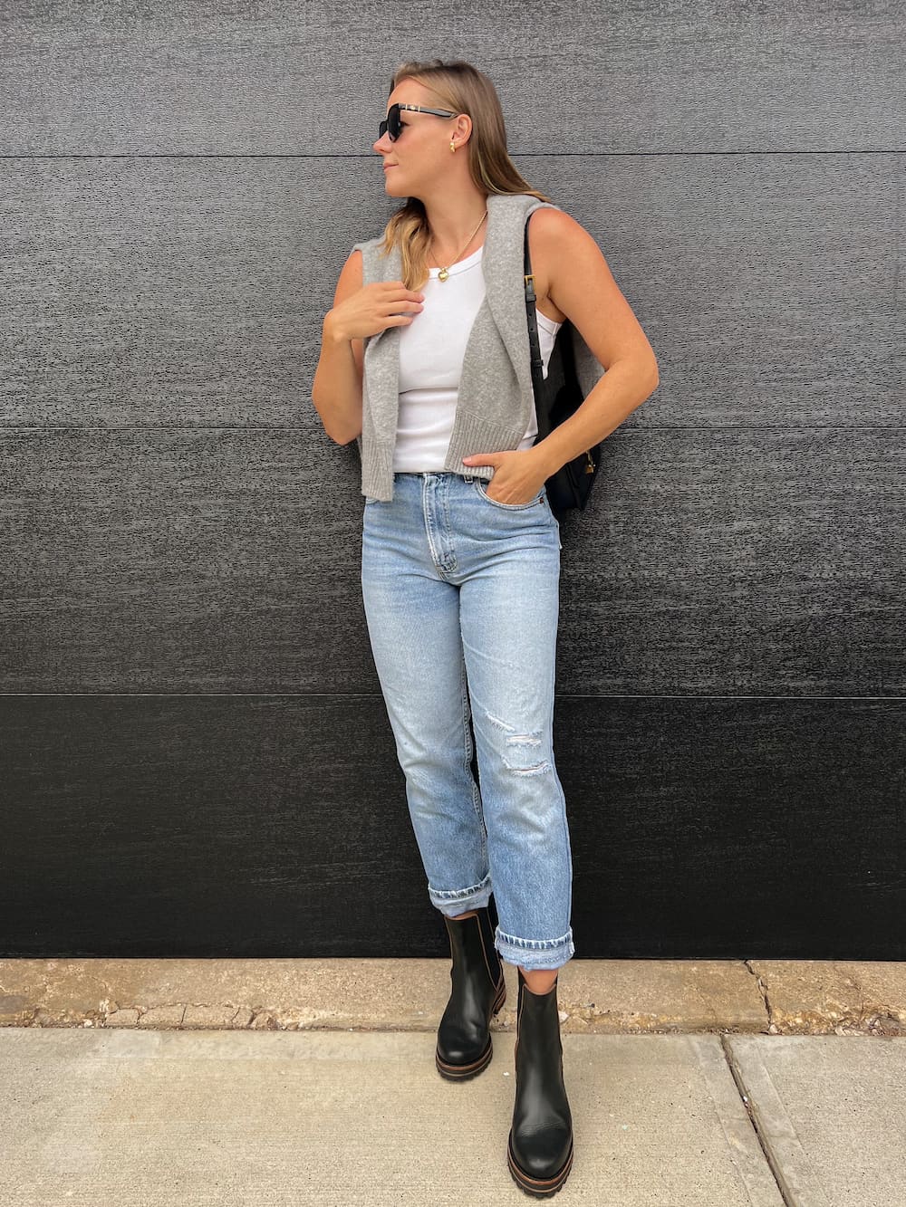Woman wearing jeans, black Chelsea boots, a white tank and a grey sweater over her shoulder.