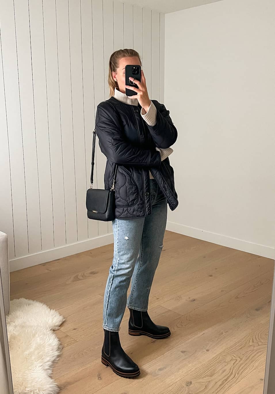 Woman wearing jeans, black chelsea boots, a white turtleneck sweater and a black quilted jacket.